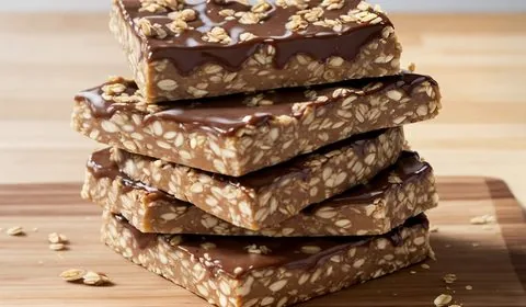 No-bake peanut butter oatmeal granola bars sit on a piece of wax paper on a dark wood plank background. Adjacent to the bars is a spoon with a scoop of peanut butter, a small glass bowl of pretzels, and a small glass bowl of oats. Oats and pretzel pieces are sprinkled about. This is the designated pinterest pin.