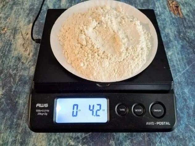 A paper plate holding flour rests on top of a weighing scale on a blue background.