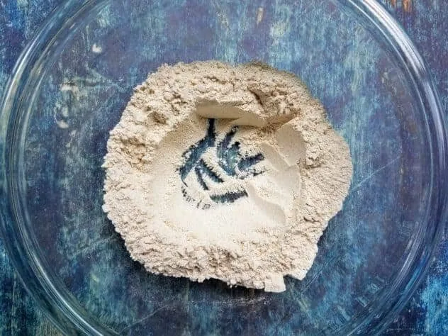 A well made by the dry ingredients in a large glass bowl on a blue background.