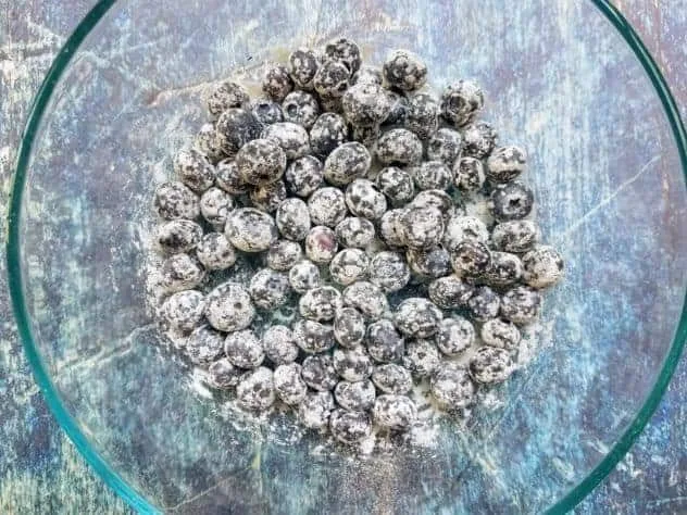 A small glass bowl on a blue background holding flour coated blueberries.