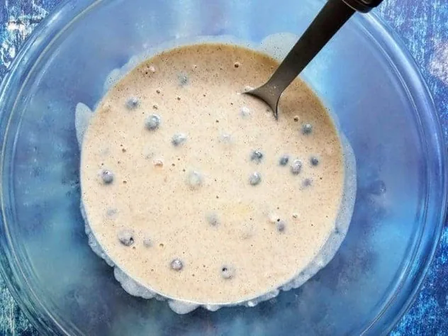 Blueberries uniformly distributed in pancake batter in a large glass bowl on a blue background.