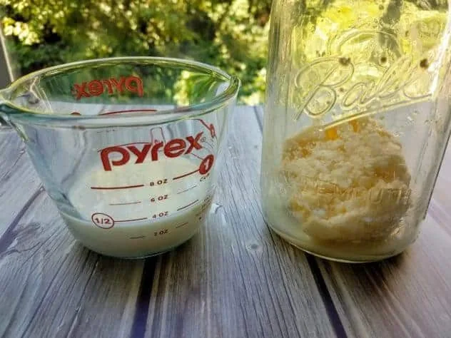 Homemade Butter Process Step 4b Buttermilk in a Pyrex measuring cup and homemade butter in a large mason jar. Both sit on a gray wood plank background.