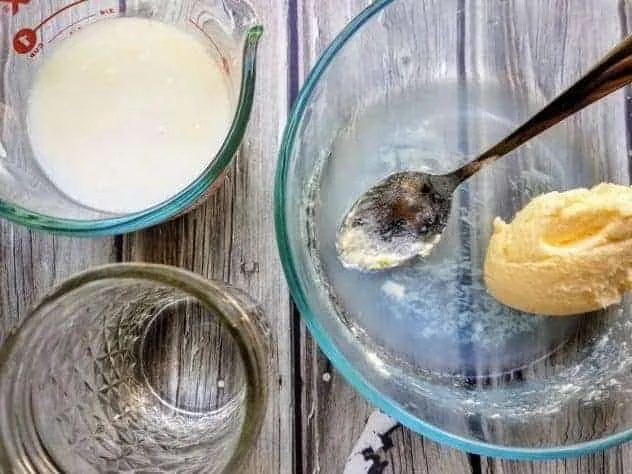 Homemade Butter Process Step 5a Buttermilk in a Pyrex measuring cup, homemade butter in a small glass bowl, and cold water in a small mason jar. All three sit on a gray wood plank background.