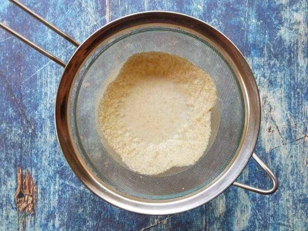 Homemade Oat Milk Process Step 4a Straining oat milk through a fine mesh stainless steel sieve over a large glass bowl on a blue background.