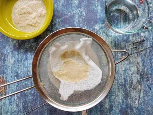 Homemade Oat Milk Process Step 4b Straining oat milk through a cheesecloth covered fine mesh stainless steel sieve over a large glass bowl on a blue background. Oat pulp sits in a green bowl in the upper left corner and 1 cup of water sits in a Pyrex measuring cup in the upper right.