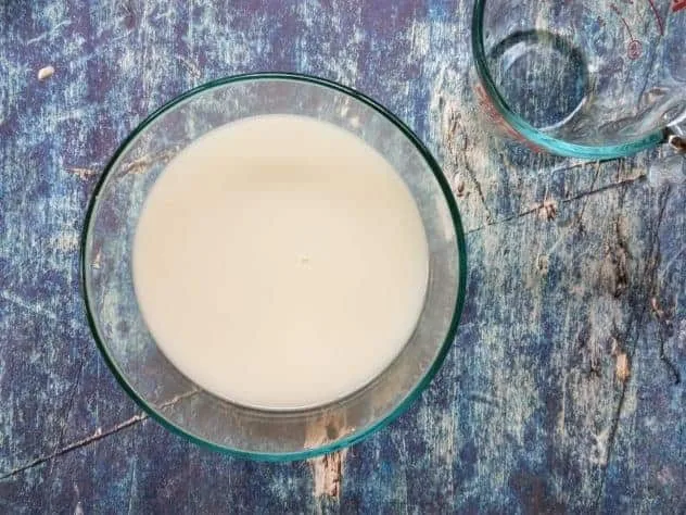 Homemade Oat Milk Process Step 5 Finished homemade oat milk sits in a large glass bowl with an empty Pyrex measuring cup in the upper right corner. Everything sits on a blue background.