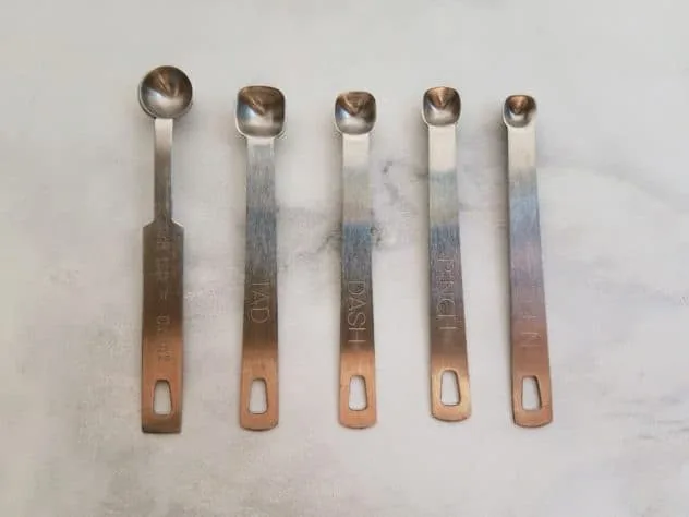 Roasted Winter Squash Seeds Process Step 2a Side-by-side comparison of the 1/8, tad, dash, pinch, and smidgen measuring spoons. Spoons sit on a white marble background.