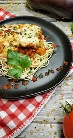 A slice of baked eggplant parmesan sits on a bed of angel hair pasta. The pasta is drizzled with tomato sauce, and garnished with a sprig of parsley. The entire thing sits on a round black plate, on top of a red/white checkerboard patterned kitchen towel. In the background is a tomato, eggplant, a sprig of basil, and some crumbs of homemade shake and bake. Peeking through the scene is also the wood plank background. This is the vertical photo.