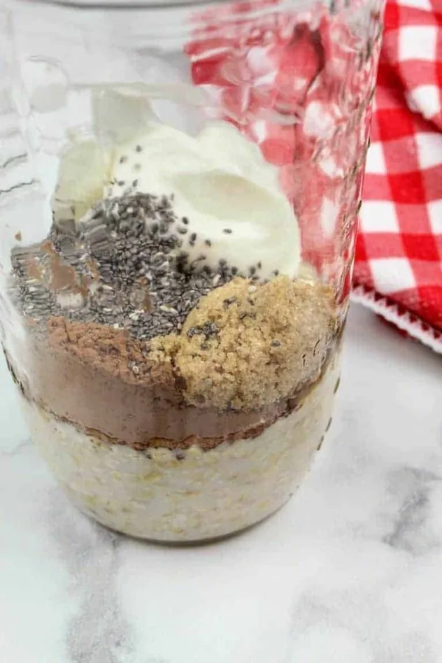 Chocolate Overnight Oats Process Step 2b Side view of chia seeds, Greek vanilla yogurt, quick oats, light brown sugar, and unsweetened cocoa powder sitting on top of the unsweetened almond milk in a glass mason jar. To the right is a white and red checkered tea towel. Everything sits on a white marble background.