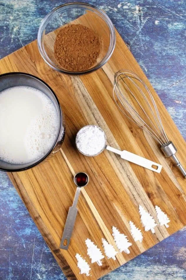 Vegan Hot Cocoa Process Step 1 Starting from the left and moving clockwise: a navy blue mug filled with almond milk that has been warmed up, a small glass bowl with cocoa powder, a small whisk, a tablespoon of confectioner's sugar, and a 1/4 teaspoon of vanilla extract all sit on a wooden cutting board. At the bottom of the cutting board are pine trees made of confectioner's sugar. The cutting board sits on a navy blue background.
