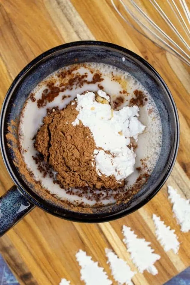 Vegan Hot Cocoa Process Step 2a Addition of cocoa powder, confectioner's sugar, and vanilla extract to the mug filled with almond milk. The whisk sits in the upper right. At the bottom of the cutting board are pine trees made of confectioner's sugar. The cutting board sits on a navy blue background.