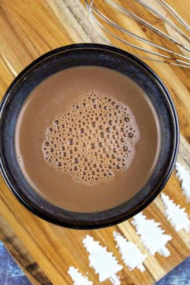 Vegan Hot Cocoa Process Step 2b Vegan hot cocoa after mixing everything in and heating it up to the desired temperature. The whisk sits in the upper right. At the bottom of the cutting board are pine trees made of confectioner's sugar. The cutting board sits on a navy blue background.