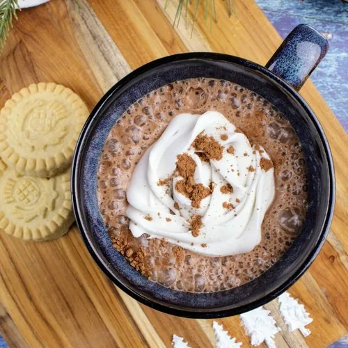 In the center is a navy colored mug filled with vegan hot cocoa. It is topped with a swirl of coconut whipped cream and cocoa powder. Off to the left are two shortbread cookies and a white tea towel. At the top is a sprig of evergreen and pine cones. In the upper right is a small dark wooden bowl of marshmallows. Everything but the marshmallows sit on a wooden cutting board, the bottom of which has a line of evergreen trees made of confectioner's sugar. Everything sits on a navy blue background. This is the vertical image.