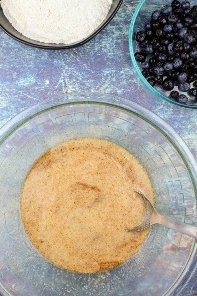 At the bottom is a large glass mixing bowl with baking powder, kosher salt, ground cinnamon, vegan butter, extra light olive oil, applesauce, and almond milk all mixed together. In the upper right is a small bowl of blueberries and in the upper left is a black bowl with gluten-free flour. Everything sits on a navy blue background.