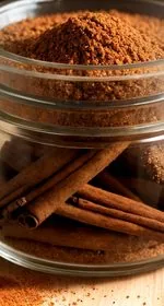 A teaspoon of apple pie spice sits in the foreground, with a jar of the spice, four apples, and two cinnamon sticks sit in the background. The jar and the teaspoon sit on a red and white checkered kitchen towel, whereas the apples and cinnamon sticks sit on a wood plank background. At the top of the image are the words 'The Panicked Foodie,' with the i's colored red and dotted with red hearts. At the end is a black mixing bowl with a red heart on it that contains three sugar cubes.