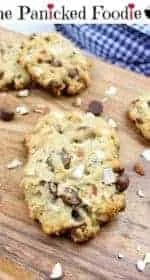 Five vegan almond coconut chocolate chip cookies sit on a dark wooden serving board. Chocolate chips, chopped almonds, and chopped coconut are scattered about on the serving board. In the upper right is a navy blue and white checkered tea towel. At the top of the image are the words 'The Panicked Foodie,' with the i's colored red and dotted with red hearts. At the end is a black mixing bowl with a red heart on it that contains three sugar cubes.