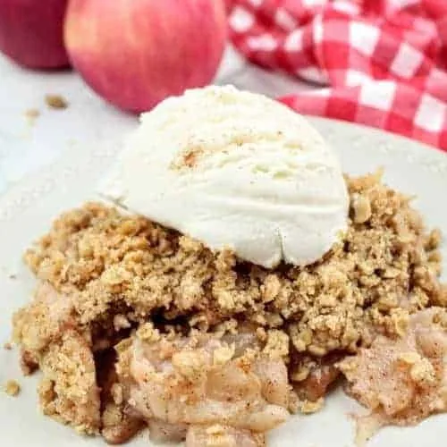 Vegan gluten-free apple crisp topped with a scoop of vanilla ice cream sprinkled with ground cinnamon sits on a white plate in the center of the photo. In the upper right is a glass pan with apple crisp and in the upper left are two red apples. Sprinkled about are crumbs of the apple crisp topping. To the right of the plate is a white and red checkered tea towel. To the left of the plate is the white marble background.